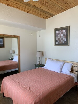 Une chambre avec un plafond en bois, un lit recouvert d’une couvre-lit à motifs roses, une table de chevet avec une lampe et une œuvre d’art encadrée accrochée au mur.