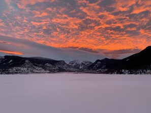 Une vue panoramique d’un paysage enneigé, avec des montagnes et un lac, sous un ciel flamboyant au coucher du soleil.