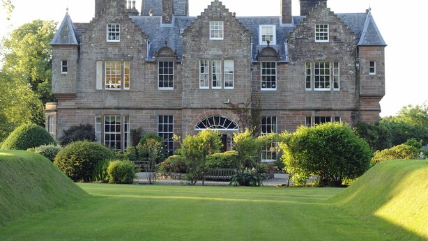 A large, historic stone building with multiple chimneys, arched windows, and a well-maintained garden. - Carnell Private Apartment (Hurllford)