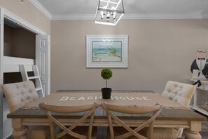 A dining area with a table labeled 'BEACHHOUSE', two upholstered chairs, a potted plant, and a framed picture of a beach scene. - Treasure Island 0205 (Panama City Beach)