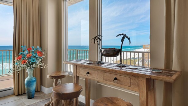 A balcony with a view of the ocean, a wooden desk with drawers, a vase of flowers, and two stools. - Treasure Island 0412 (Panama City Beach)