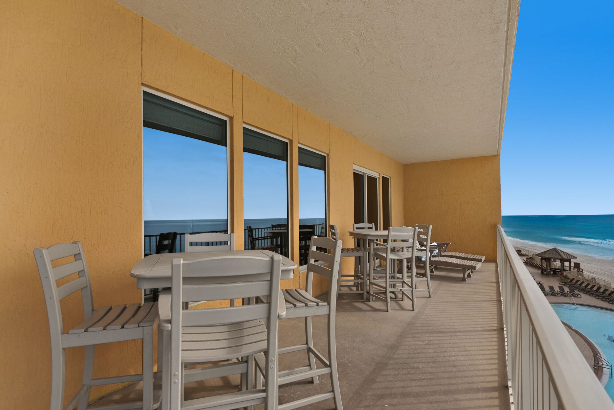 A balcony with white chairs and tables, overlooking a beach and a pool.