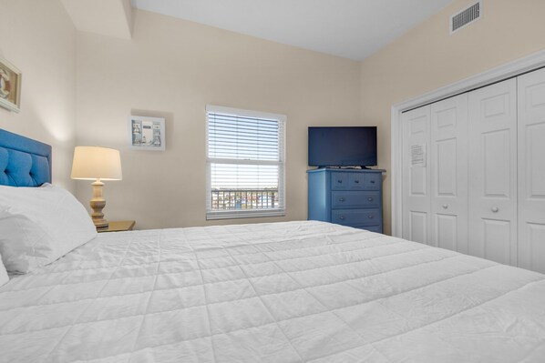 A bedroom with a bed, a nightstand, a lamp, a window with blinds, a dresser, a television, and a wardrobe.