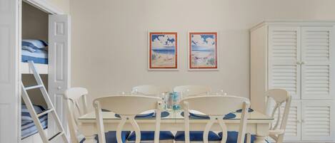 Zona pranzo con tavolo apparecchiato per quattro, circondato da sedie bianche con cuscini blu. In evidenza due quadri appesi alla parete, un armadio bianco e una scala che conduce a un letto a castello.