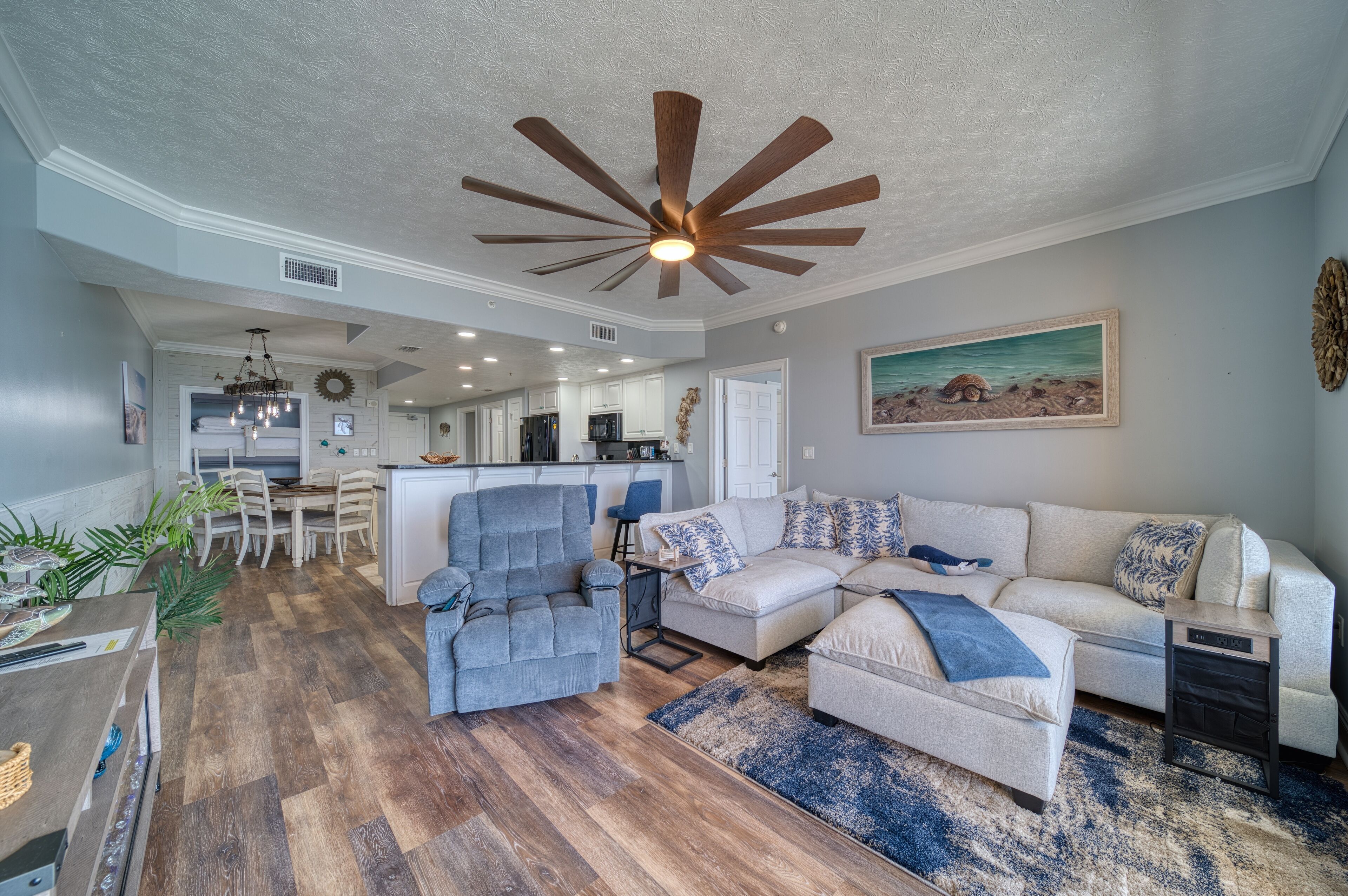 A spacious living room with a ceiling fan, a sofa set, a dining area, and a kitchen in the background.