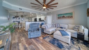 A spacious living room with a ceiling fan, a sofa set, a dining area, and a kitchen in the background.