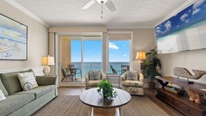 A living room with a ceiling fan, a green sofa, a coffee table with a plant, a wooden sideboard, and a large painting of a beach. - Treasure Island 1009 (Panama City Beach)