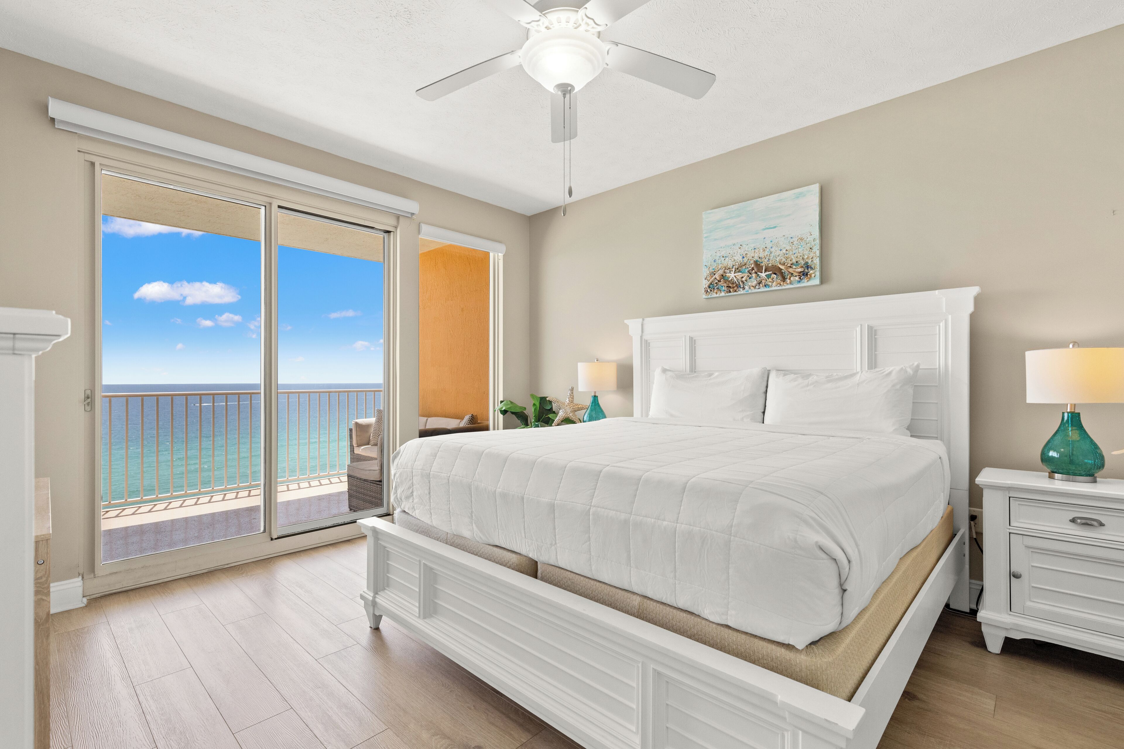 A bedroom with a bed, a ceiling fan, a nightstand with a lamp, and a view of the ocean through a sliding door.