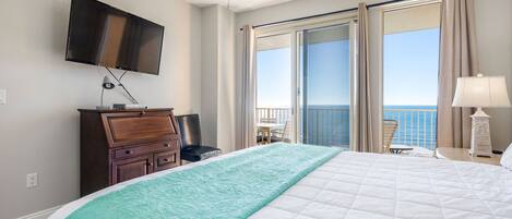 A hotel room with a large bed, a TV mounted on the wall, a desk with a chair, and a balcony with a view of the ocean.