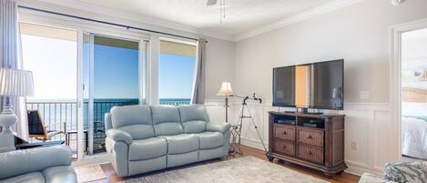 A living room with a sofa, a television, and a balcony with a view of the ocean.