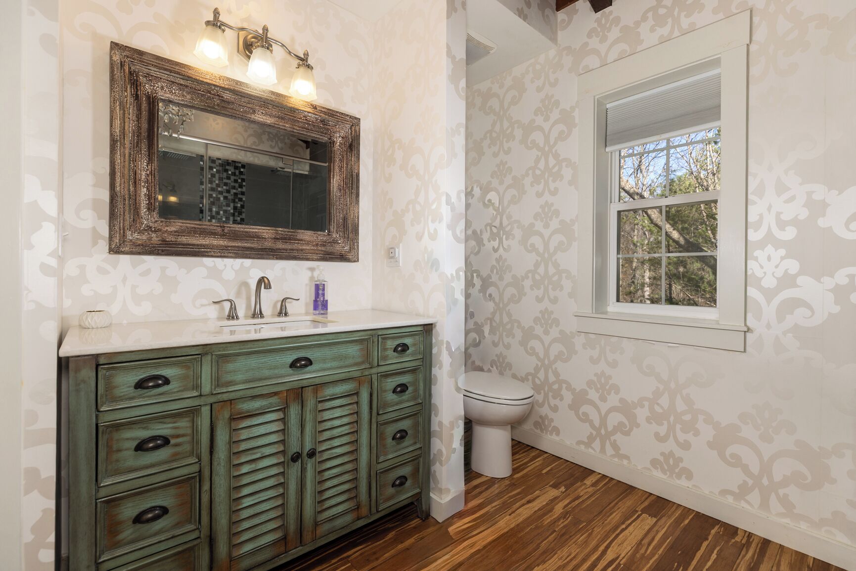 Un baño con una tocador de estilo vintage, un espejo grande y una ventana con vistas a árboles.