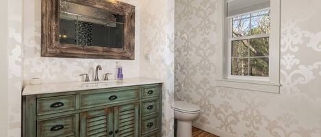 Un baño con una tocador de estilo vintage, un espejo grande y una ventana con vistas a árboles.