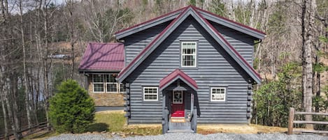 A log cabin with a red roof, a front porch, and a gravel driveway.