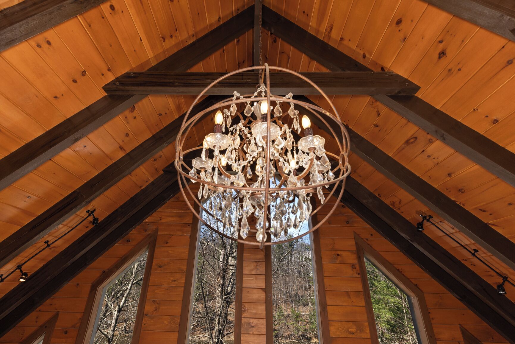 A wooden interior with a large chandelier, wooden beams, and windows with a view of trees.