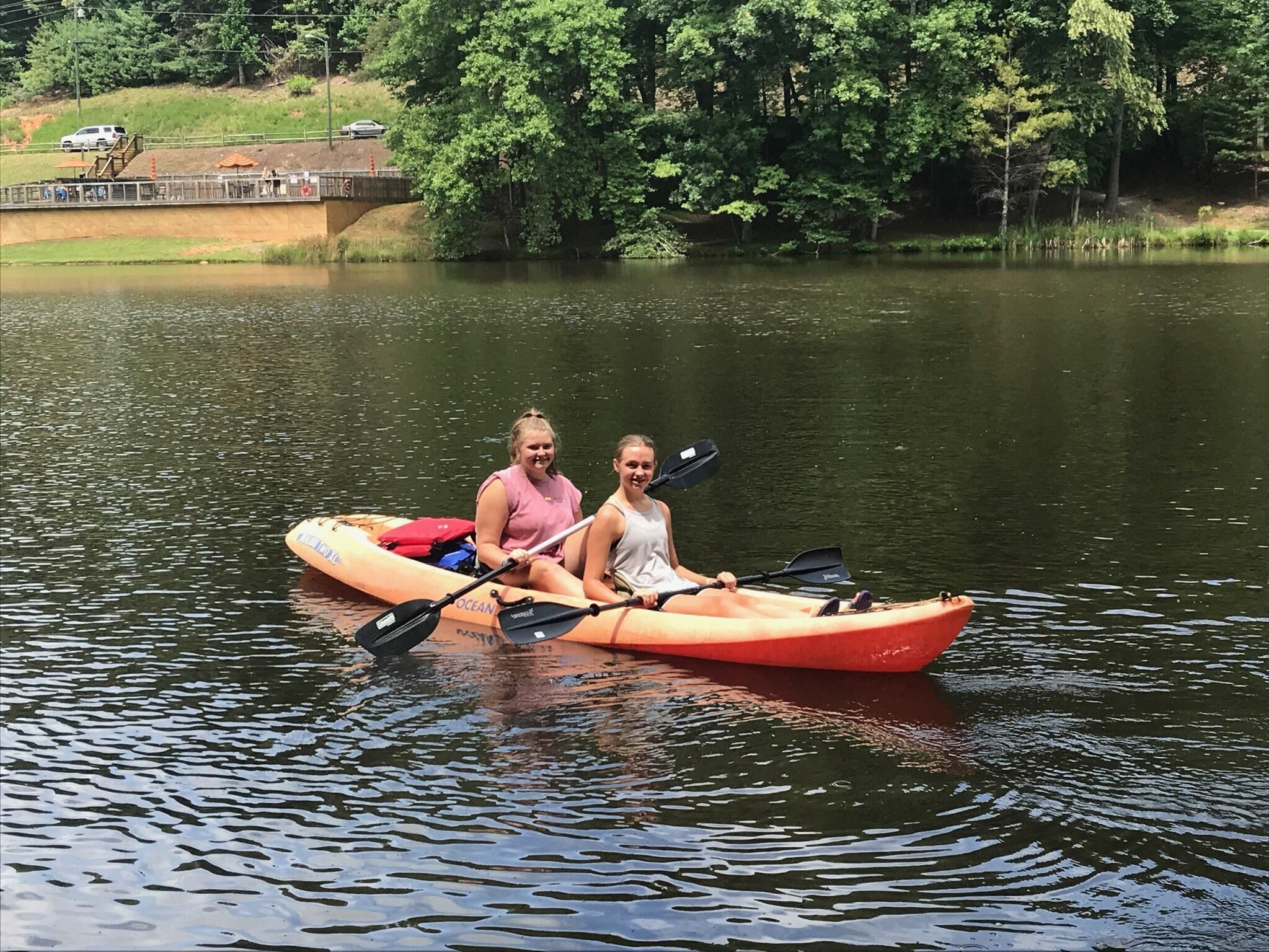 Two people in a red kayak on a calm lake with a bridge and trees in the background.