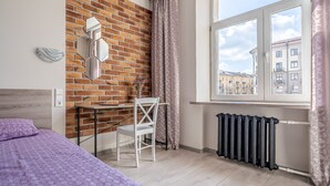 Una camera da letto con parete in mattoni, una finestra vista edifici, un termosifone, un letto con copriletto fantasia, una scrivania con sedia e un elemento decorativo a parete.