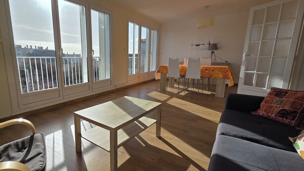 Ein geräumiges Wohnzimmer mit Holzboden, einem grauen Sofa, einem kleinen Tisch und einem Balkon mit Blick auf Gebäude.