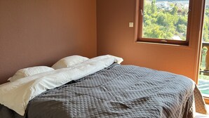 Un dormitorio con cama, paredes marrones y ventana con vistas a zonas verdes y edificios.