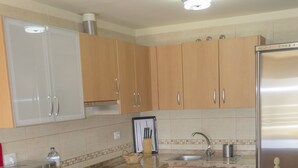 A kitchen with wooden cabinets, a stainless steel refrigerator, a microwave, a sink, and a bowl of fruit on the counter. There are three white bar stools in front of the counter.