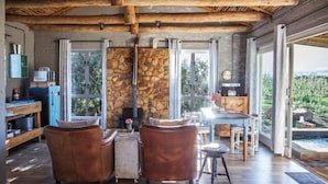 A rustic interior with stone walls, wooden beams, leather chairs, a stone fireplace, and large windows offering a view of the outdoors. - Bergsicht Country Farm Cottages (Tulbagh)
