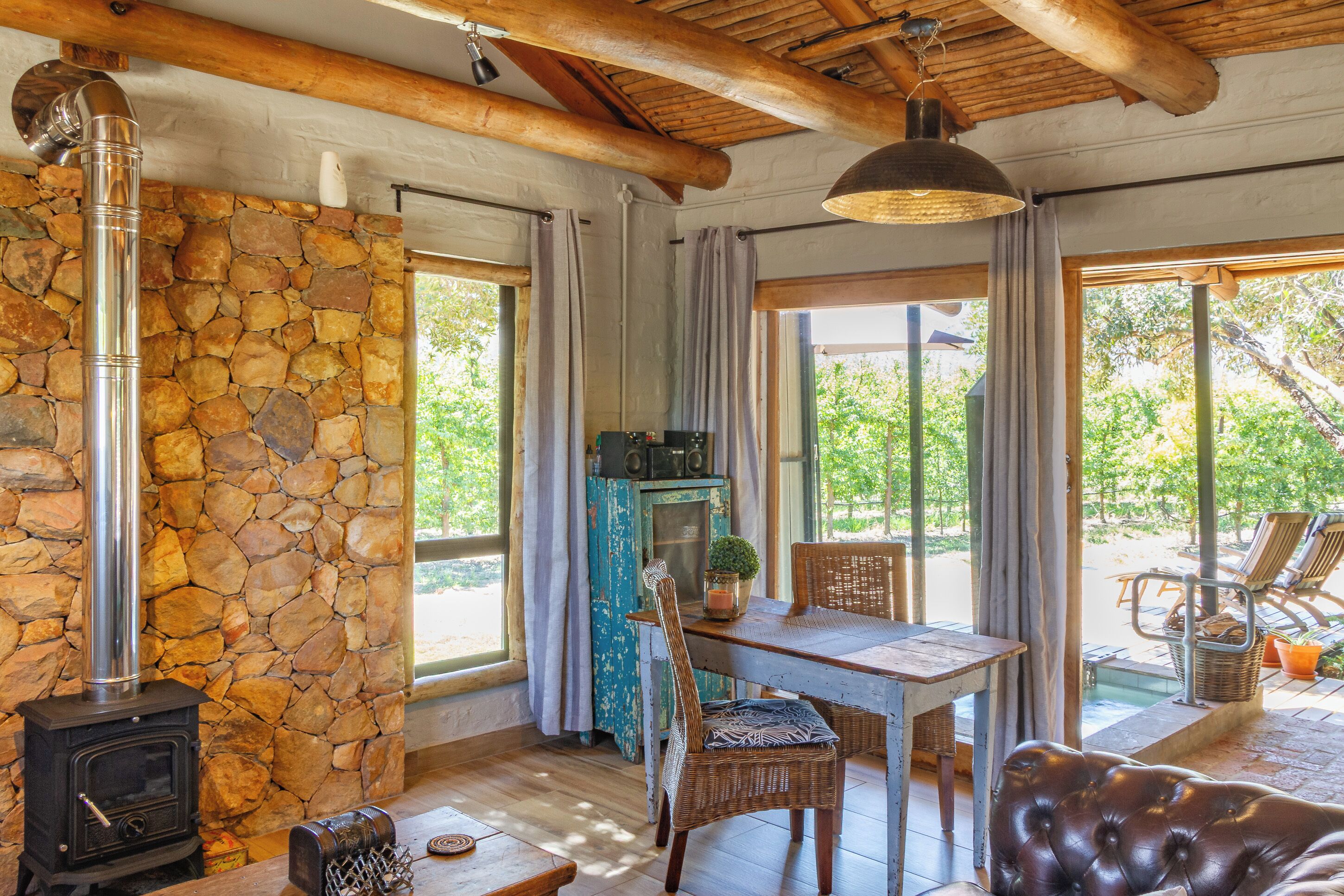 A rustic interior with a stone wall, a wood-burning stove, a dining table with chairs, and a view of the outdoors through large windows.