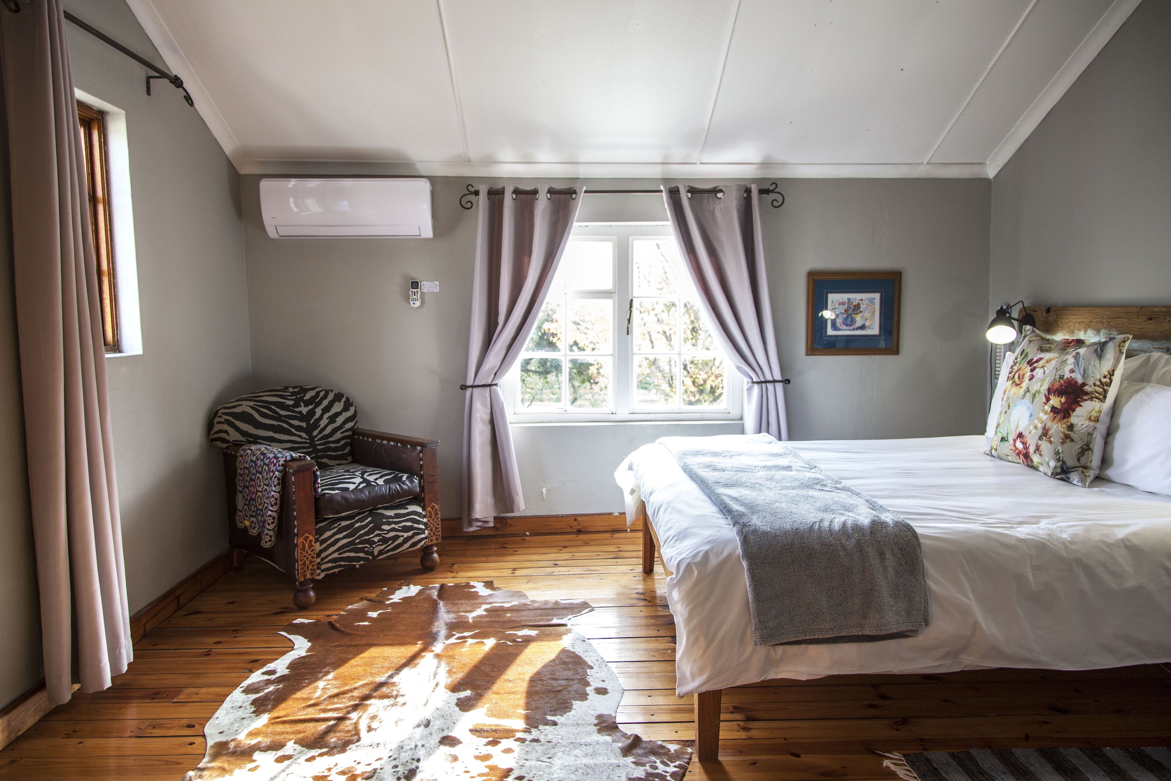 Un dormitorio con suelo de madera, una cama con ropa de cama blanca, una silla con estampado de cebra, una alfombra de cuero de vaca y una ventana con cortinas.