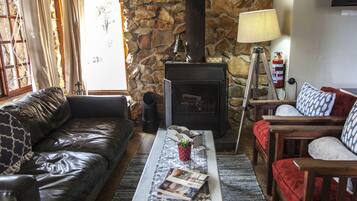 A cozy living room with a stone wall, leather sofa, wooden chairs, a fireplace, and a coffee table with magazines.