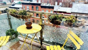 Un balcón con una mesa y sillas amarillas, plantas en macetas y vistas a edificios.