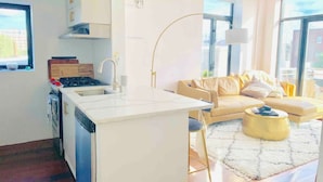 Una cocina moderna con una isla central, una zona de comedor con sofá y mesa de centro, y amplias ventanas con vistas a la ciudad.