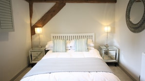 A neatly made bed with white bedding and striped pillows, flanked by two bedside tables with lamps, in a room with exposed wooden beams and a round mirror on the wall.