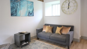 A living room with a grey sofa, a side table, a large abstract painting, a round wall clock, and a window.
