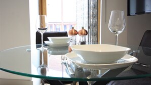 Une table à manger avec un plateau en verre, des assiettes blanches, des bols, des couverts et deux verres à vin.