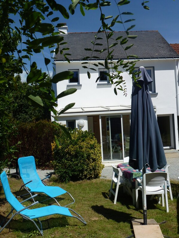 A white house with a grey roof, a patio with blue chairs, and a table with a tablecloth.