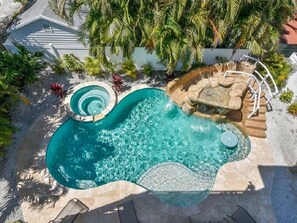 A swimming pool with a hot tub, surrounded by palm trees and a stone deck.