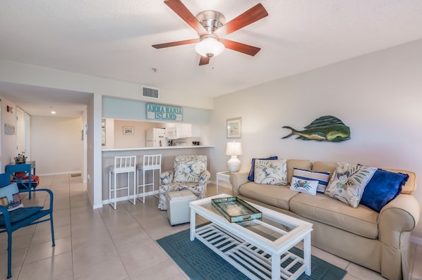 A living room with a beige sofa, a coffee table, and a ceiling fan. There is a kitchen area with a bar and a sign that reads "Anna Maria Island".