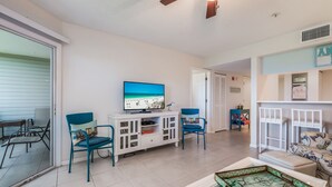 Ein Wohnzimmer mit Fernseher, der eine Strandansicht zeigt, zwei blauen Sesseln, einem weißen Schrank und einem Glastisch.