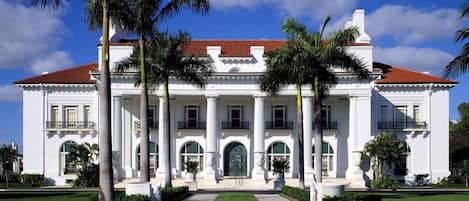 A large white building with a red-tiled roof, surrounded by palm trees and a well-maintained lawn.