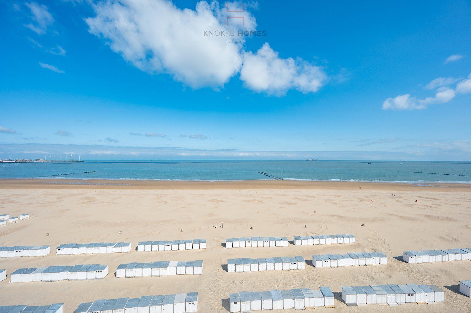 Une plage bordée de rangées de petits chalets blancs aux toits bleus, un ciel bleu dégagé et une mer calme à l’horizon.
