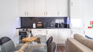 A modern kitchen with white cabinets, a black countertop, and stainless steel appliances. There is a dining table with chairs and a sofa in the living area. - Savory Beige Apartment, AC, Wifi !New! (Lisboa)
