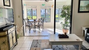 A living room with a TV, a dining table with chairs, a coffee table, and a sliding glass door leading to a patio.