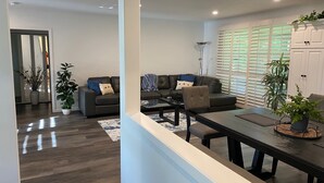Una sala de estar moderna con un sofá, sillones y una mesa de comedor. Hay plantas en macetas y un ventanal con persianas.