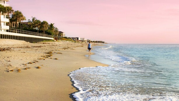 A beach with a person walking along the shore, palm trees, and buildings in the background. - Ready to Rest and Relax at Vero Villa- Your Vacation Home Awaits. (Vero Beach)