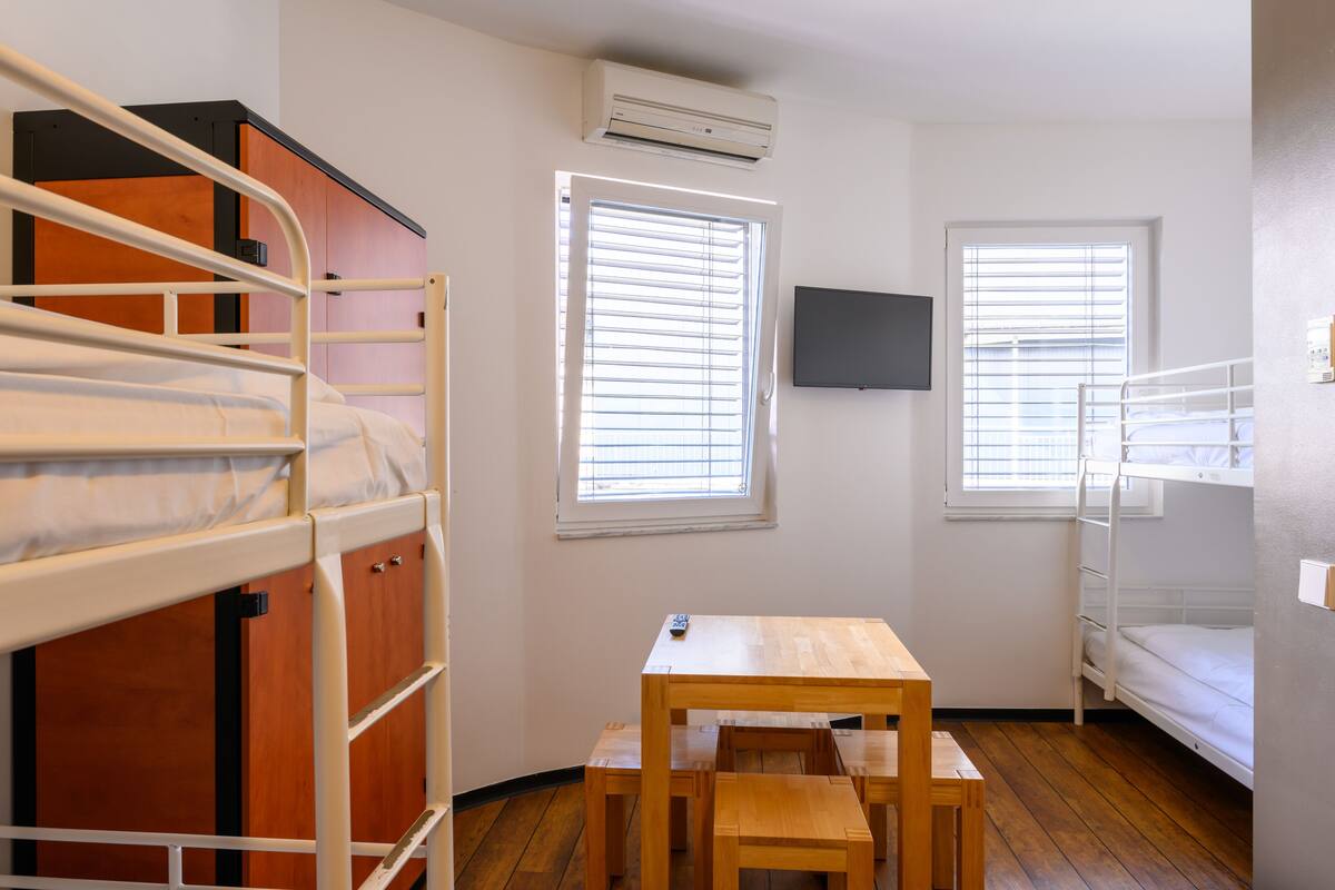 Une chambre de dortoir avec des lits superposés, une table en bois et deux fenêtres équipées de stores.