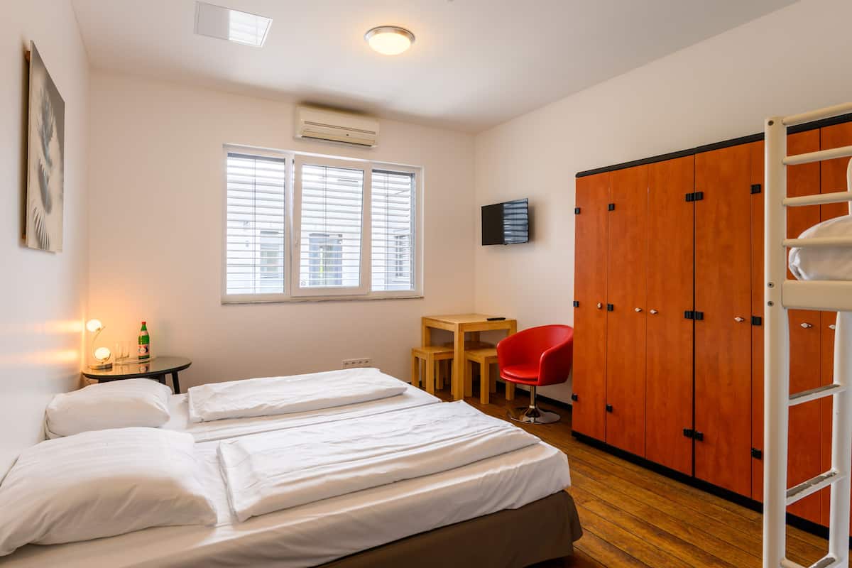 Une chambre d’hôtel avec deux lits, un bureau, une chaise rouge, une armoire et une fenêtre avec des stores.