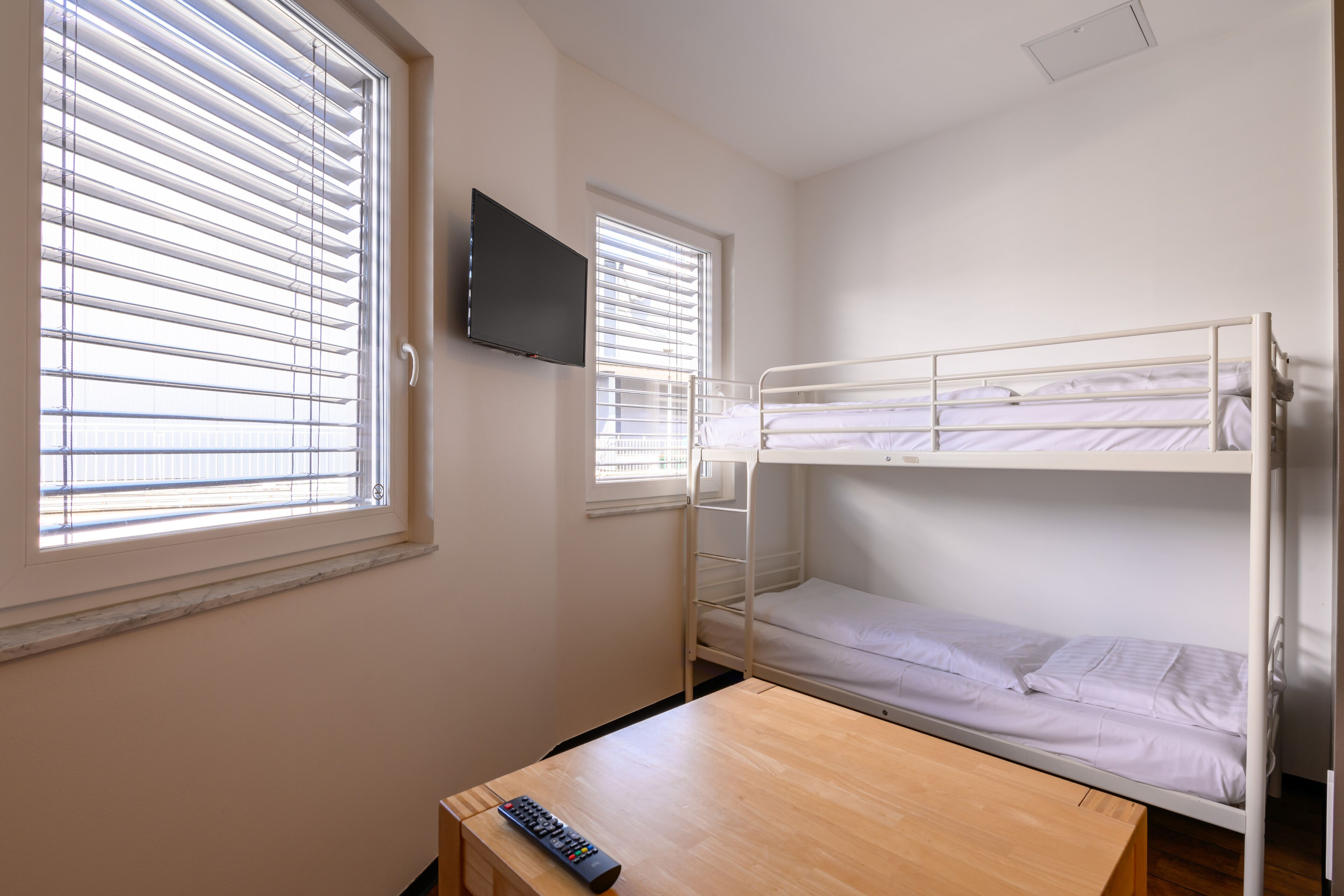 A bunk bed room with a TV on the wall, a wooden table, and two windows with blinds.