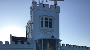 Une tour blanche, surmontée d’une tourelle et dotée d’une grande fenêtre, est entourée d’un mur blanc et d’une barrière faite de chaînes.