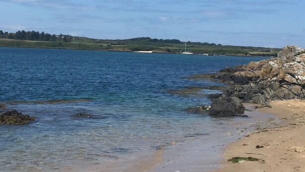 Une plage à l’eau d’un bleu limpide, avec un rivage rocheux et des collines qui se dessinent à l’horizon.