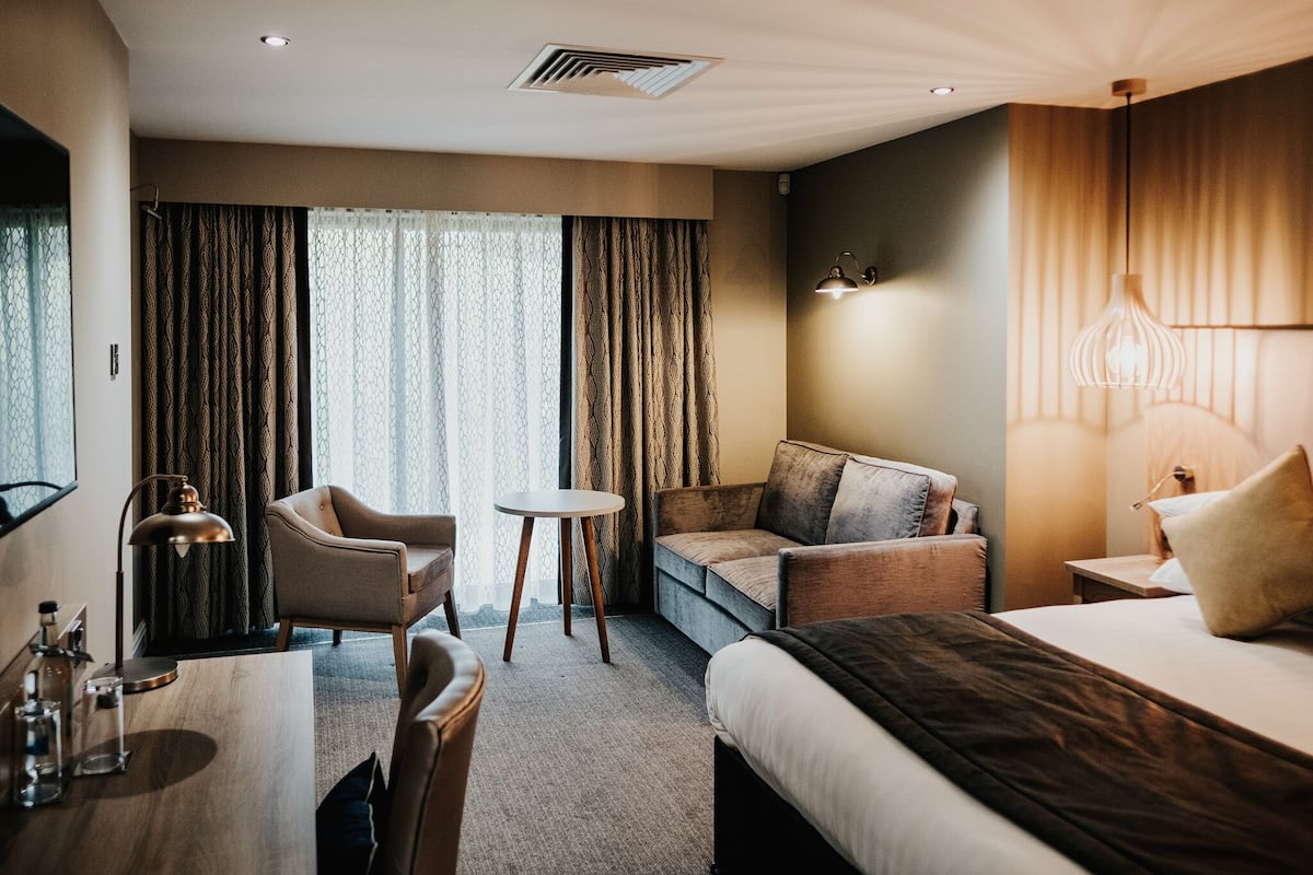 Une chambre d’hôtel avec un lit, un canapé, une chaise, une petite table et une lampe.