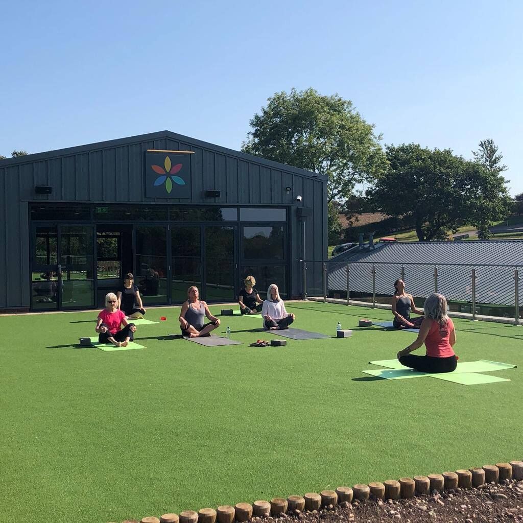 A group of people sitting on yoga mats outdoors in front of a building with a logo on the wall.