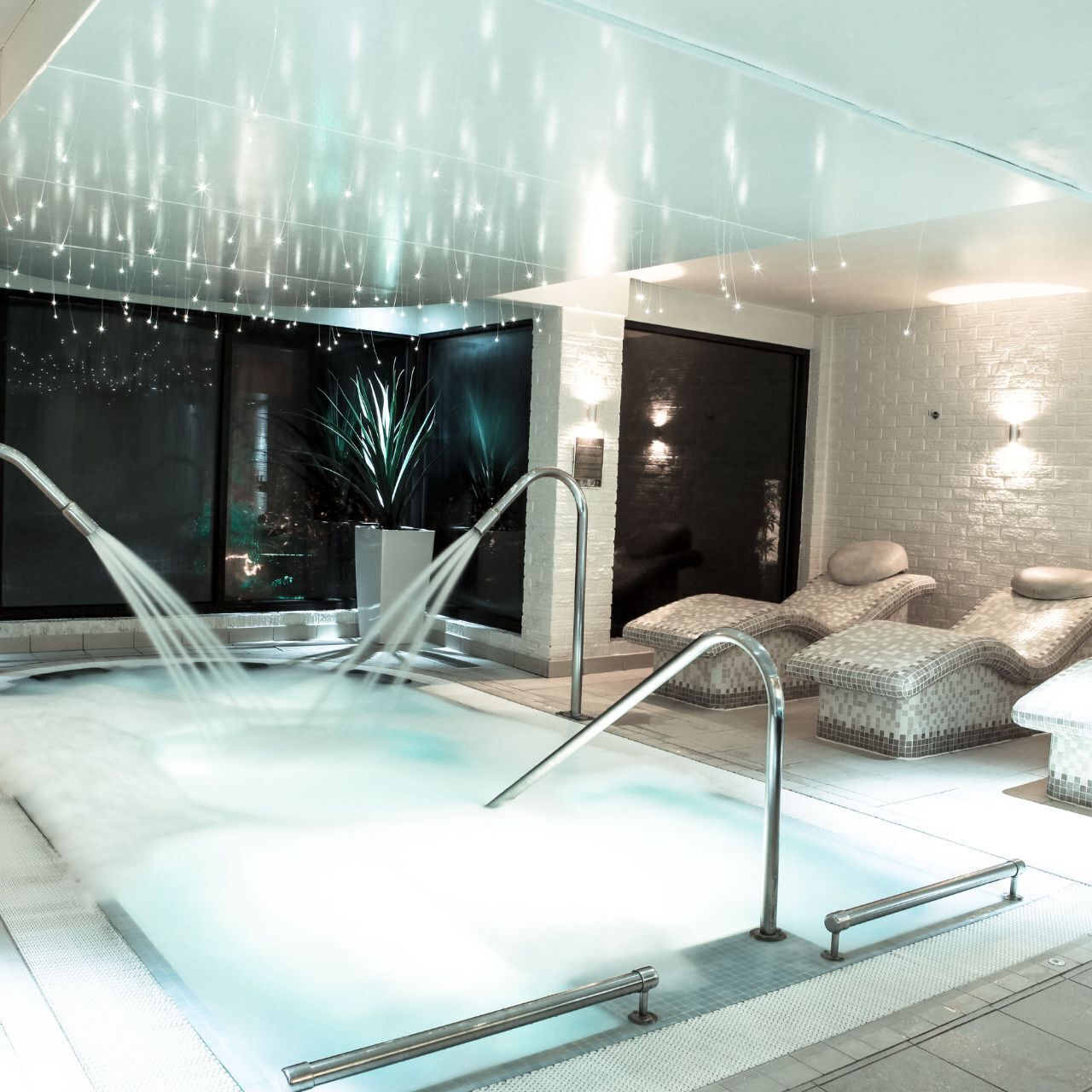 A modern indoor pool area with a jacuzzi, seating area, and a view of the cityscape at night.